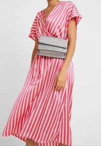 Borsa crossbody glitter argento con tre scomparti impilati e una sottile tracolla in catena, esposta su un vestito a righe rosa e bianche.