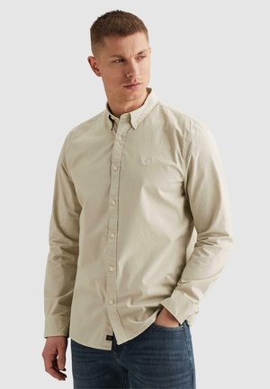 Young man with short hair wearing a beige button-up shirt and blue jeans, looking to his right with one hand in his pocket.