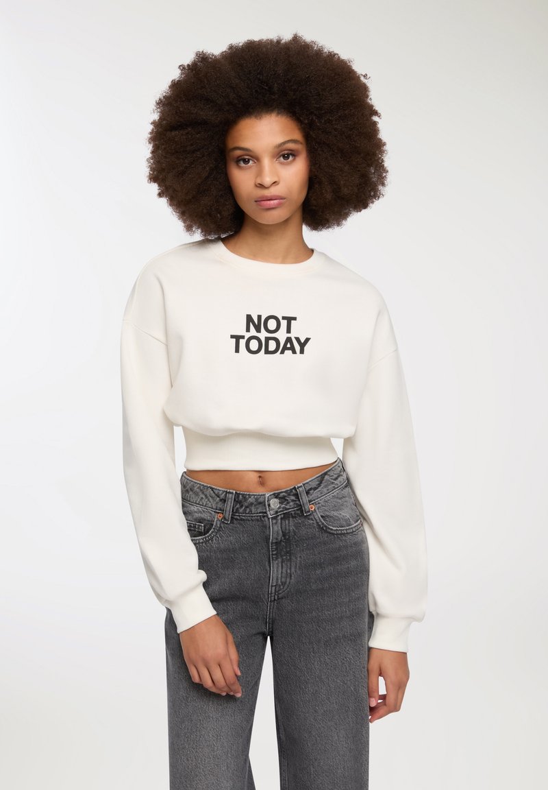 Felpa bianca cropped con testo nero "NOT TODAY", caratterizzata da maniche ampie e un orlo aderente, abbinata a jeans grigi a vita alta.