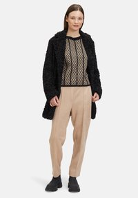 Schwarzer strukturierter Mantel, beiger Rippenpullover mit schwarzem geometrischem Muster, beiger weit geschnittener Hose und schwarzen Knöchelstiefeln.