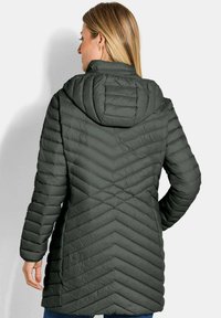 Gevoerde lange jas in donkergroen met een chevron-quilt ontwerp. Voorzien van een capuchon en lange mouwen, gemaakt van een lichtgewicht materiaal.
