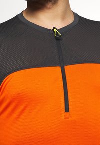 Camiseta atlética de manga corta en negro y naranja con textura de malla, cierre de medio zip y detalle reflectante cerca del cuello.