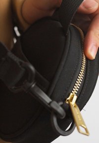 Juodas audinio maišelis su auksine užtrauktuko ir aparatūros detalėmis, turintis apvalią formą ir tekstūruotą dirželį. Artima nuotrauka, kurioje matyti ranka laikanti maišelį.