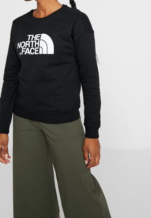 Person trägt schwarzen Pullover mit großem weißem "The North Face"-Logo und olivgrünen, weitem Hosenbein vor einfachem Hintergrund.