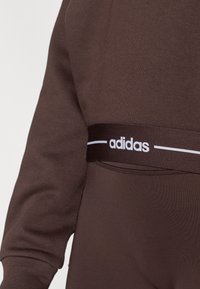 Pruun spordiriietus, mis koosneb dressipluusist ja retuusidest, millel on must vöökoht valge "adidas" logoga ning sile tekstuur.