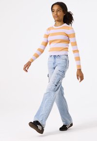 Top de manga larga a rayas en naranja y lila, combinado con unos vaqueros cargo azul claros con múltiples bolsillos y un corte relajado. Zapatillas negras.