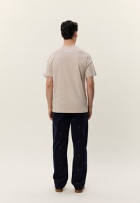 Férfi, aki bézs rövid ujjú inget és mintás fekete nadrágot visel, háttal áll egy egyszerű világos háttér előtt.