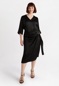 Black satin dress with a V-neck, three-quarter sleeves, and a wrap design. Includes a gold circular accent at the waist. Strappy heels.