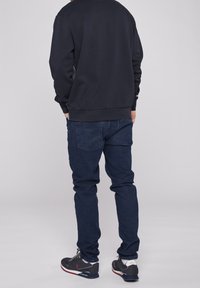 Marinblå crewneck-tröja, jeans och grå sneakers. Outfiten är avslappnad med en ledig passform och synliga bakfickdetaljer.