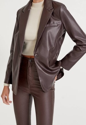Person wearing a brown leather blazer and matching fitted pants with a cream ribbed turtleneck crop top, one hand in pocket.