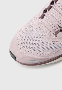 Αθλητικό παπούτσι σε ανοιχτό ροζ χρώμα με διαπνέον επάνω μέρος από πλέγμα, υφασμάτινες λεπτομέρειες και διακριτικές αντανακλαστικές πινελιές. Η σόλα είναι σκοτεινή με λεία επιφάνεια.