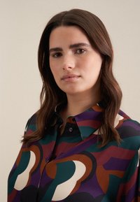 Camicia con maniche lunghe e bottoni, caratterizzata da audaci motivi geometrici in verde acqua, bordeaux, panna e viola. Tessuto liscio con colletto e bottoni neri.