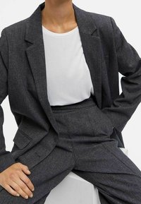 Costume gris ajusté en tissu texturé, comprenant un blazer à revers et un pantalon taille haute, associé à une chemise blanche à col rond.