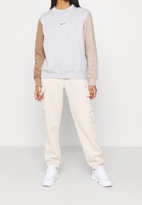 Nike sweatshirt with gray body and beige sleeves, featuring a small logo. Paired with cream cargo pants and white sneakers, on a plain backdrop.