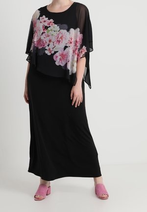 Personne portant une longue robe noire avec une surcouche florale transparente ornée de fleurs roses, associée à des chaussures roses ouvertes, debout devant un fond uni.