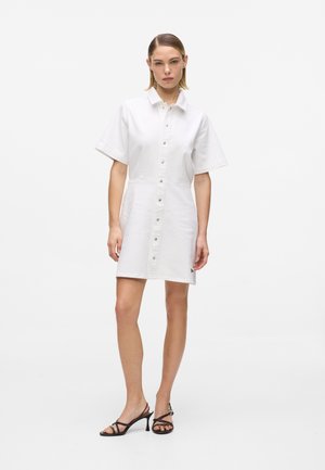 Γυναίκα που στέκεται φορώντας λευκό φόρεμα με κουμπιά και κοντά μανίκια και μαύρα σανδάλια με τακούνι και λουράκια σε απλό λευκό φόντο.