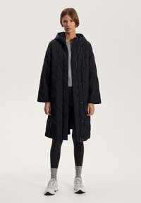 Manteau long en tissu matelassé noir avec une capuche, des fermetures à pression et des fentes sur les côtés. Porté sur un haut gris et des leggings noirs, présentants une texture lisse.