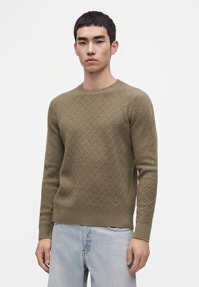 Giovane uomo che indossa un maglione verde oliva lavorato a girocollo e jeans azzurri, in piedi davanti a uno sfondo bianco semplice.