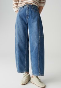 Jean large bleu clair en denim avec une taille haute, deux poches avant, et des détails de couture subtils. Associé à des baskets beiges.