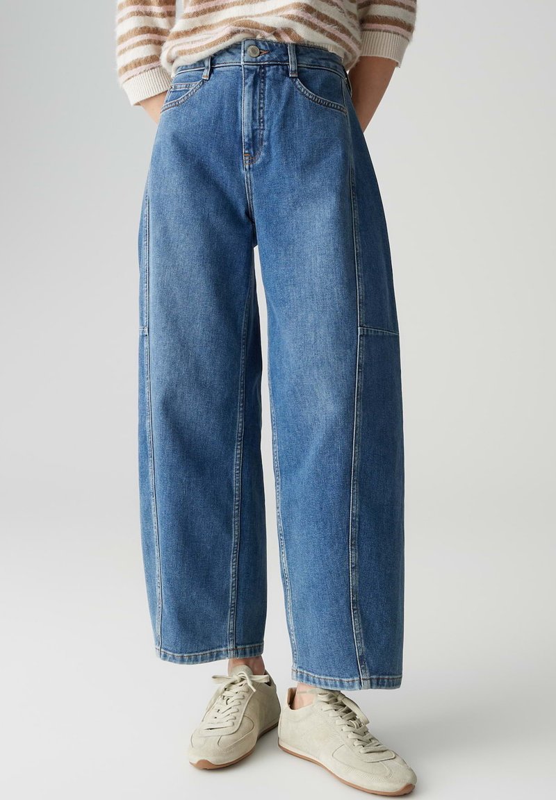 Jean large bleu clair en denim avec une taille haute, deux poches avant, et des détails de couture subtils. Associé à des baskets beiges.