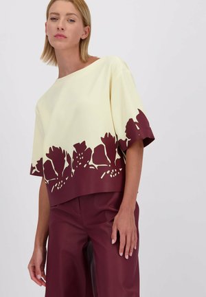 Moderne cropped top in lichtgeel met een maroon bloemen uitsnede aan de onderrand; heeft een losse pasvorm en korte mouwen.