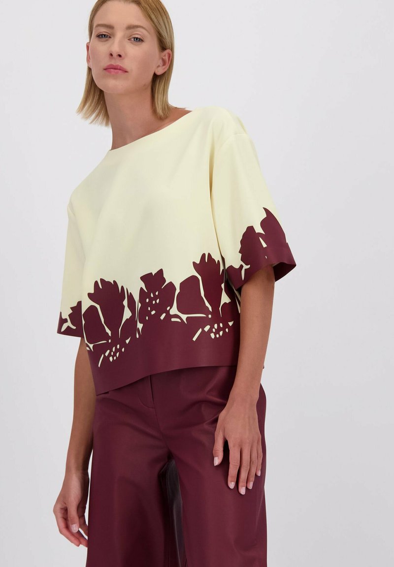 Moderne cropped top in lichtgeel met een maroon bloemen uitsnede aan de onderrand; heeft een losse pasvorm en korte mouwen.