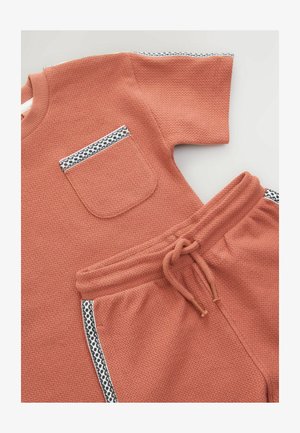 Terakotas divdaļīgs apģērbs, kas sastāv no teksturēta īsām piedurknēm topa ar krūšu kabatu un atbilstošām biksēm ar rakstainiem sānu akcentiem.