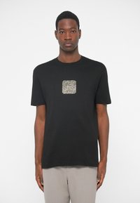 Camiseta preta com um design texturizado em forma de quadrado de cor metálica ao centro. Tem mangas curtas e um decote redondo.