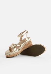 Goldene metallische Keilsandalen mit mehreren überkreuzten Riemen, Kork-ähnlicher Sohle und einem strukturierten Fußbett für zusätzliche Stabilität.