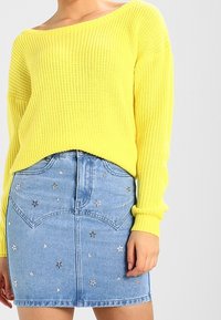 Gelber gestrickter Pullover mit weitem Ausschnitt, kombiniert mit einem hellblauen Jeansrock, der mit silbernen Sternapplikationen verziert ist und eine taillierte Silhouette hat.