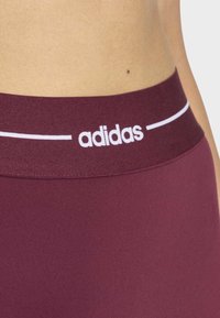 Cinturilla atlética color granate con la marca "adidas" en blanco y una línea horizontal de acento; textura suave y diseño ajustado para ropa deportiva.