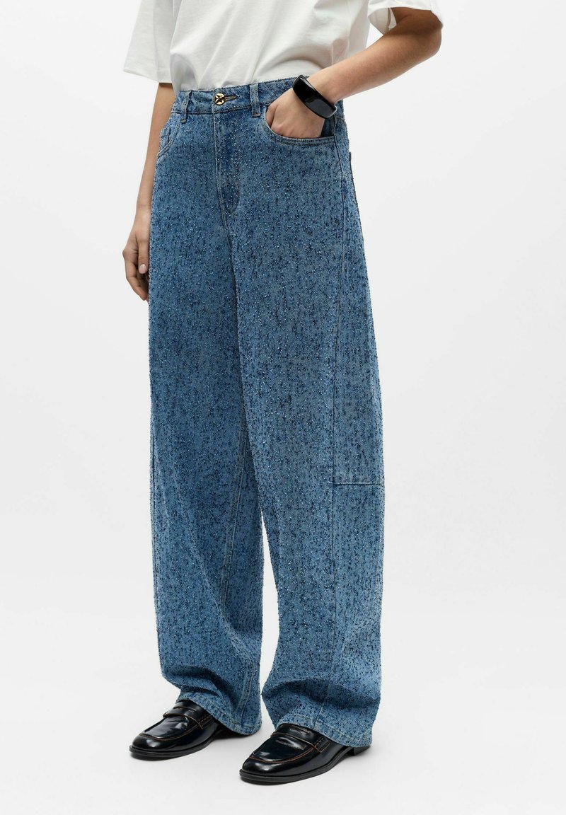 Personne portant un jean bleu ample à motifs mouchetés, une chemise blanche à manches courtes, un bracelet noir épais et des mocassins en cuir noirs, debout avec une main dans la poche.