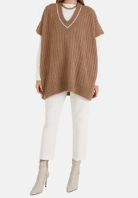 Brązowy, prążkowany, luźny sweter bez rękawów z dekoltem w serek i kontrastującą białą lamówką, zestawiony z białymi spodniami i beżowymi botkami za kostkę.
