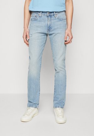 Ανοιχτόμπλε τζιν με ίσια εφαρμογή, ελαφρά φθορά στο γόνατο και σχέδιο πέντε τσέπες, σε συνδυασμό με λευκά αθλητικά παπούτσια.