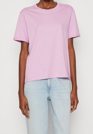 Osoba ubrana w jednolity, jasnofioletowy T-shirt z krótkim rękawem oraz jasnoniebieskie dżinsy, stojąca na tle jednolitego białego tła.