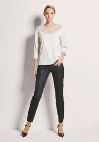 Roomkleurige top met kanten inzetstuk, driekwartmouwen; gecombineerd met zwarte slim-fit broek en bordeauxrode hoge hakken. Neutrale achtergrond.