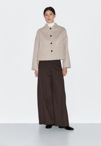 Vrouw in een beige cropped knopenjack, witte coltrui, brede donkere bruine broek en zwarte schoenen, staand tegen een witte achtergrond.