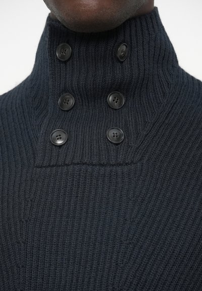 Tummansininen ribattu neulepaita korkealla kauluksella, jossa on neljä mustaa nappia ja tekstuuri, joka tuo lisäyksityiskohtia.