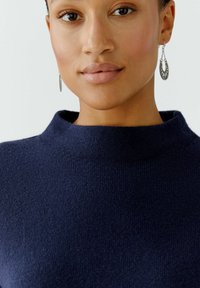 Dunkelblauer Pullover mit hohem Kragen, aus einem weichen, strukturierten Stoff. Silberne Creolen ergänzen den Look.