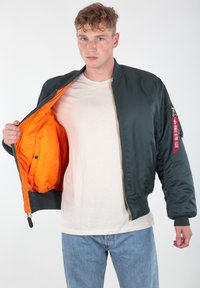 Zelena bomber jakna s narančastim oblogom, zatvaranje na patentni zatvarač, rebrasti rukavi i crvena oznaka na rukavu. Glatki, lagani materijal.