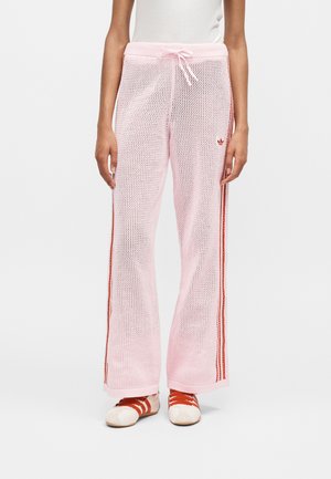 Személy világos rózsaszín hálós, bő szárú nadrágot visel, oldalán piros csíkokkal, és fehér sportcipőt piros fűzővel.