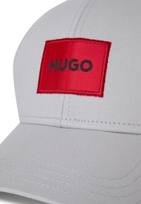Gorra gris con un parche rectangular rojo que presenta la palabra "HUGO" en negro. Fabricada en un tejido suave, con forma estructurada y un notable detalle de costura.