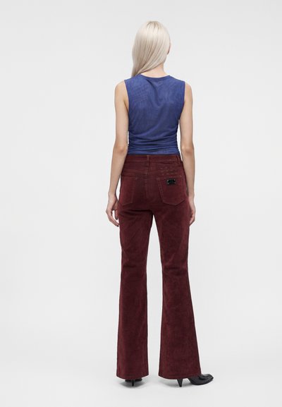 Femme aux longs cheveux blonds, de dos, vêtue d'un haut bleu sans manches, un pantalon évasé bordeaux et des chaussures noires à bouts pointus.