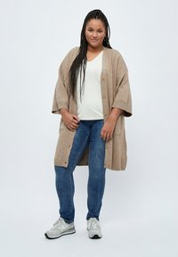 Cardigan beige oversize con chiusura a bottoni, indossato sopra una t-shirt bianca a scollo a V, abbinato a jeans skinny blu e sneakers grigie.
