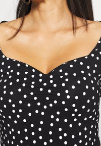 Svart topp med V-ringning och vita prickar. Tillverkad av stretchigt material med samlad detalj vid bysten.