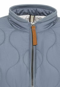 Himmelblau gesteppte Jacke mit Stehkragen, schwarzem Reißverschluss und einem braunen Leder-Zugriemen. Sie weist dezente, geschwungene Nähmuster auf.
