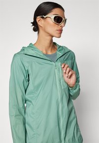 Lightweight, hooded jacket in soft mint green fabric, features a full zipper and a zippered chest pocket. Accompanied by stylish sunglasses.