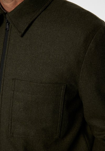 Olivgrön jacka i texturerad tyg med full dragkedja och en ficka på framsidan för funktionell detalj.