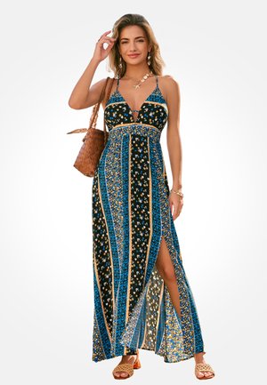 Vrouw draagt een mouwloze maxi-jurk met bloemenprint en een zijslit, ze draagt een gevlochten bruine tas en accessoires van een schelpenketting en oorbellen.