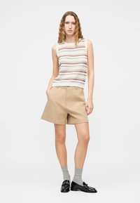 Γυναίκα που φοράει ριγέ πλεκτό τοπ χωρίς μανίκια, μπεζ σορτς, γκρι κάλτσες και μαύρα μοκασίνια, στέκεται μπροστά σε απλό λευκό φόντο.